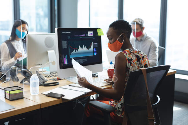 Африканская американка в маске для лица читает документ, сидя на столе в современном офисе. социальная изоляция от карантина во время пандемии коронавируса — стоковое фото