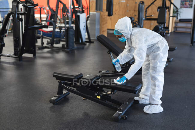 Travailleuse portant des vêtements de protection et un masque facial nettoyant la salle de gym à l'aide de désinfectant. isolement social mise en quarantaine pendant une pandémie de coronavirus — Photo de stock