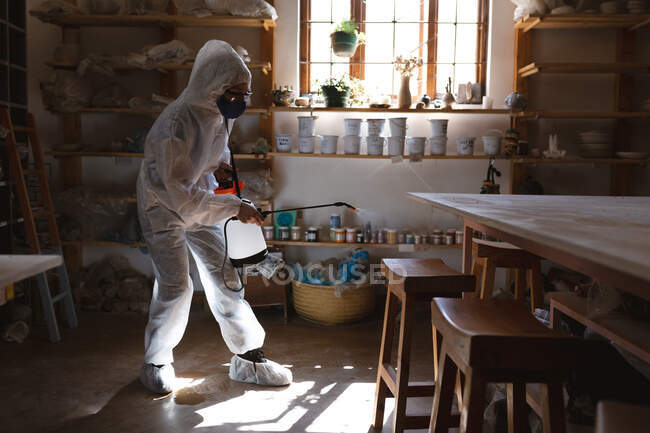 Nettoyant masculin caucasien en vêtements de protection travaillant dans un atelier de poterie. désinfecter tout l'endroit. petite entreprise créative pendant la pandémie de coronavirus covid 19. — Photo de stock