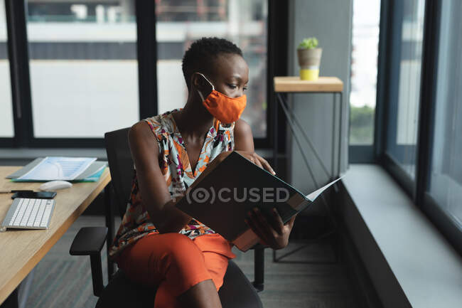Mujer afroamericana con mascarilla en la oficina. sentado en el escritorio leyendo documentos. higiene y distanciamiento social en el lugar de trabajo durante la pandemia de coronavirus covid 19. - foto de stock
