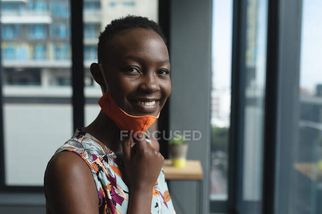 Портрет афро-американської жінки, яка тягне вниз маску обличчя і посміхається в офісі. гігієна і соціальна дистанція на робочому місці під час коронавірусу covid 19 пандемії. — стокове фото