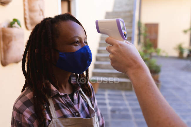 Mixed race female potter in face mask outside of pottery studio. wearing apron, having temperature checked. small creative business during covid 19 coronavirus pandemic. — Stock Photo