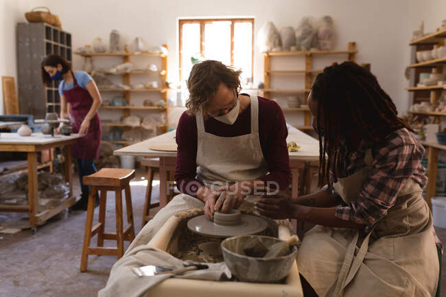 Кавказские мужчины и женщины-гончары смешанной расы в масках, работающие в мастерской керамики. в фартуках, работая на гончарном круге. малый творческий бизнес во время пандемии коронавируса ковида 19. — стоковое фото