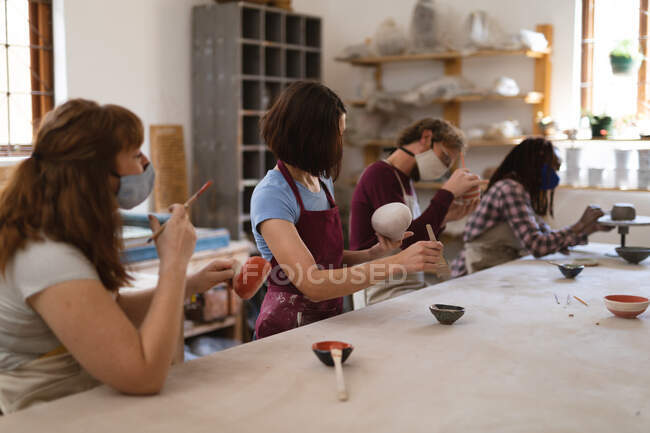 Multi-ethnic group of potters in face masks working in pottery studio. wearing aprons, painting plates. small creative business during covid 19 coronavirus pandemic. — Stock Photo