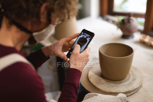 Caucasian male potter in face mask working in pottery studio. wearing apron, taking a photo of a bowl. small creative business during covid 19 coronavirus pandemic. — Stock Photo