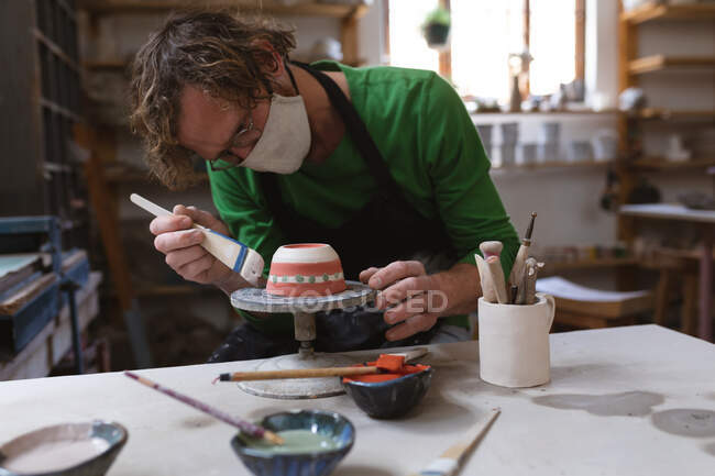 Caucasian male potter in face mask working in pottery studio. wearing apron, working at a potters wheel, painting bowl. small creative business during covid 19 coronavirus pandemic. — Stock Photo