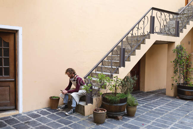 Caucasian male potter sitting on stairs outside pottery studio. wearing apron, using smartphone. small creative business during covid 19 coronavirus pandemic. — Stock Photo