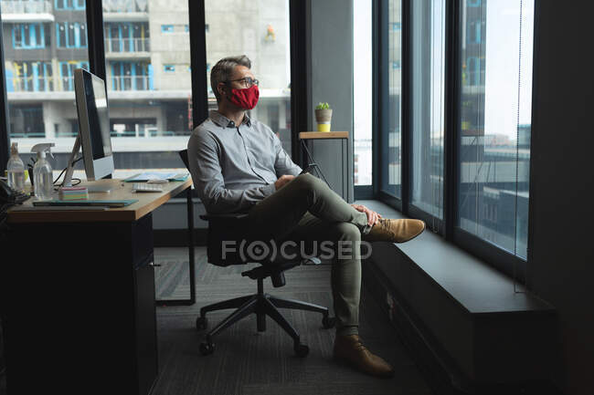 Hombre caucásico con máscara facial sentado mirando por la ventana en la oficina. sentado en el escritorio sosteniendo teléfono inteligente. higiene y distanciamiento social en el lugar de trabajo durante la pandemia de coronavirus covid 19. - foto de stock