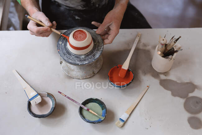 Alfarero masculino trabajando en un estudio de cerámica. trabajando en una rueda de alfareros, pintando un tazón. pequeña empresa creativa durante la pandemia de coronavirus covid 19. - foto de stock