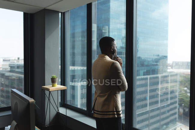 Afrikanischer Mann mit Gesichtsmaske, der aus dem Fenster schaut. in einem modernen Bürogebäude mit Blick auf die Stadt. Hygiene und soziale Distanzierung am Arbeitsplatz während der Coronavirus-Pandemie 19. — Stockfoto