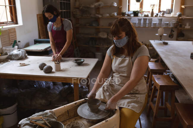 Deux femmes potiers caucasiennes masquées travaillant dans un atelier de poterie. portant un tablier, travaillant à une roue de potiers et à une table de travail. petite entreprise créative pendant la pandémie de coronavirus covid 19. — Photo de stock