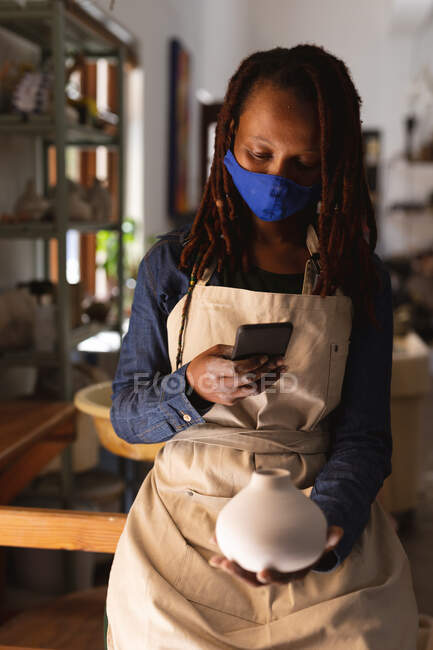 Potier mixte féminin en masque facial travaillant dans un atelier de poterie. portant un tablier, utilisant un smartphone. petite entreprise créative pendant la pandémie de coronavirus covid 19. — Photo de stock