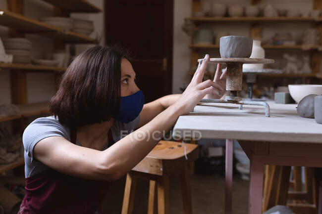 Ceramista caucasica in maschera facciale che lavora in studio di ceramica. indossando un grembiule, lavorando alla ruota di un vasaio. piccola attività creativa durante covid 19 coronavirus pandemia. — Foto stock
