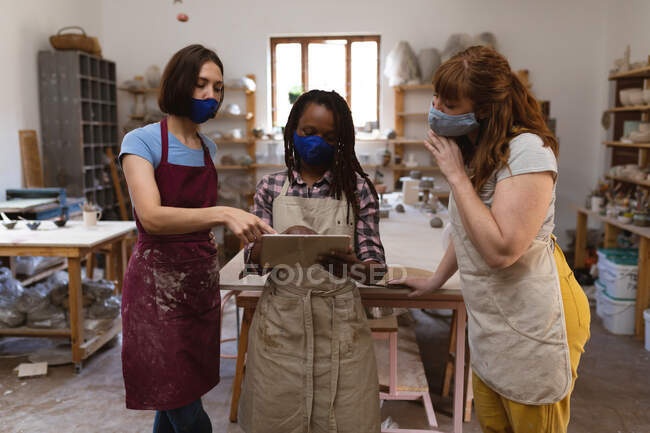 Deux potiers caucasiens et une femme métisse en masque facial travaillant dans un atelier de poterie. portant des tabliers, regardant la tablette numérique. petite entreprise créative pendant la pandémie de coronavirus covid 19. — Photo de stock