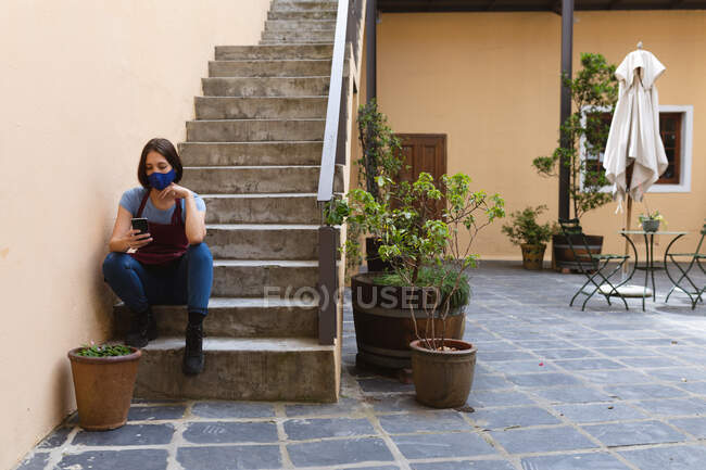 Ceramiche caucasiche sedute sulle scale fuori dallo studio di ceramica. indossando grembiule, utilizzando smartphone. piccola attività creativa durante covid 19 coronavirus pandemia. — Foto stock