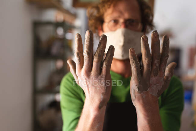 Cerâmica caucasiana macho em máscara facial trabalhando em estúdio de cerâmica. mostrando suas mãos sujas para a câmera. pequeno negócio criativo durante a pandemia do coronavírus covid 19. — Fotografia de Stock