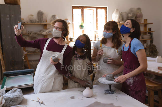 Groupe multi-ethnique de potiers masqués travaillant dans un atelier de poterie. portant des tabliers, peignant des assiettes, prenant un selfie ensemble. petite entreprise créative pendant la pandémie de coronavirus covid 19. — Photo de stock