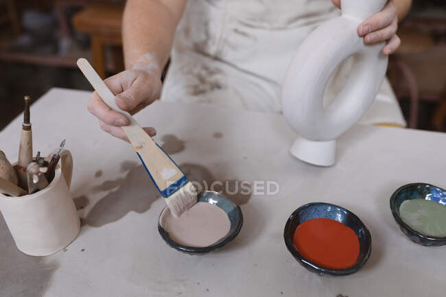 Ceramista donna che lavora nello studio di ceramica. lavorando a un tavolo di lavoro, dipingendo un piatto. piccola attività creativa durante covid 19 coronavirus pandemia. — Foto stock