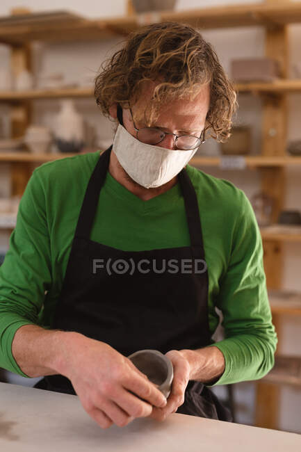 Retrato de un hombre caucásico usando mascarilla en un estudio de cerámica. pequeña empresa creativa durante la pandemia de coronavirus covid 19. - foto de stock