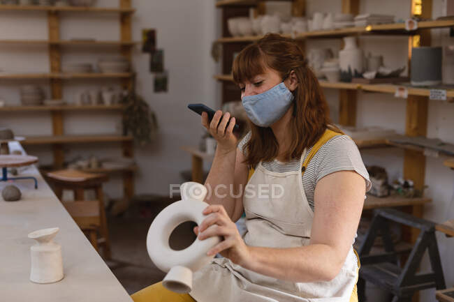 Кавказька жінка-гончар в масці обличчя працює в гончарній студії. в фартусі, розмовляючи по телефону. маленьке творче підприємство під час 19-ї коронавірусної пандемії. — стокове фото