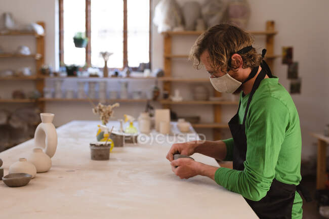 Potier masculin caucasien en masque facial travaillant dans un atelier de poterie. portant un tablier, travaillant à une table de travail. petite entreprise créative pendant la pandémie de coronavirus covid 19. — Photo de stock