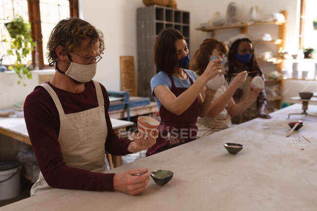 Grupo multi-étnico de oleiros em máscaras faciais trabalhando no estúdio de cerâmica. a usar aventais, a pintar pratos. pequeno negócio criativo durante a pandemia do coronavírus covid 19. — Fotografia de Stock