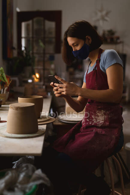 Potier femme caucasienne en masque facial travaillant dans un atelier de poterie. portant un tablier, prenant une photo d'un bol. petite entreprise créative pendant la pandémie de coronavirus covid 19. — Photo de stock