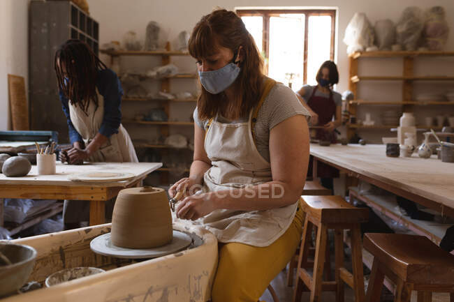 Кавказька жінка-гончар в масці обличчя працює в гончарній студії. в фартусі, працюючи на кермі з двома її друзями позаду. маленьке творче підприємство під час 19-ї коронавірусної пандемії — стокове фото