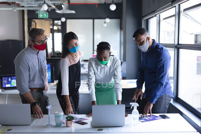 Различные коллеги носят маски для лица, используя ноутбук, работая вместе в современном офисе. гигиена и социальное дистанцирование на рабочем месте во время пандемии коронавируса 19. — стоковое фото