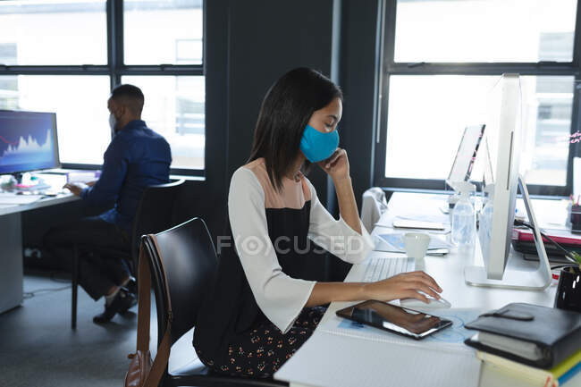 Азиатка в маске с помощью компьютера сидит на столе в современном офисе. гигиена и социальное дистанцирование на рабочем месте во время пандемии коронавируса 19. — стоковое фото