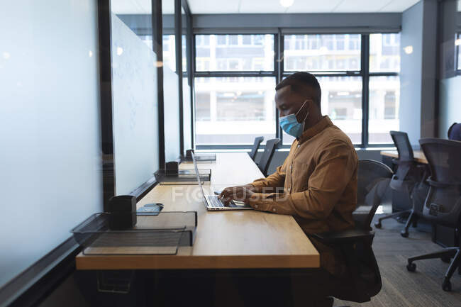 Homme afro-américain portant un masque facial à l'aide d'un ordinateur portable tout en étant assis sur son bureau au bureau moderne. isolement social mise en quarantaine pendant une pandémie de coronavirus — Photo de stock