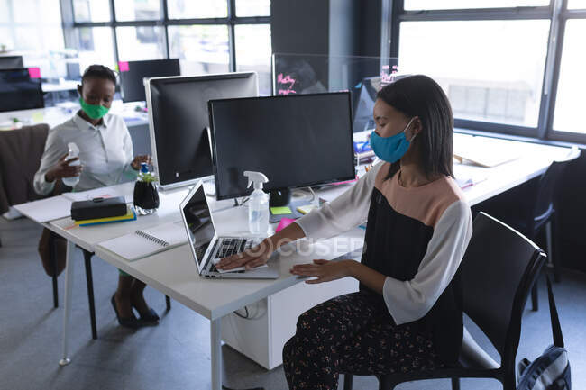 Mulher asiática usando máscara facial limpando seu laptop no escritório moderno. bloqueio de quarentena por distanciamento social durante a pandemia do coronavírus — Fotografia de Stock