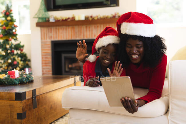 Afro-americana mãe e filha ter vídeo chat no tablet vestindo chapéus de Papai Noel durante o Natal em casa distanciamento social durante covid 19 coronavírus quarentena lockdown. — Fotografia de Stock