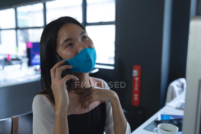 Asian woman wearing face mask talking on smartphone while sitting on her desk at modern office. hygiene and social distancing in the workplace during coronavirus covid 19 pandemic. — Stock Photo