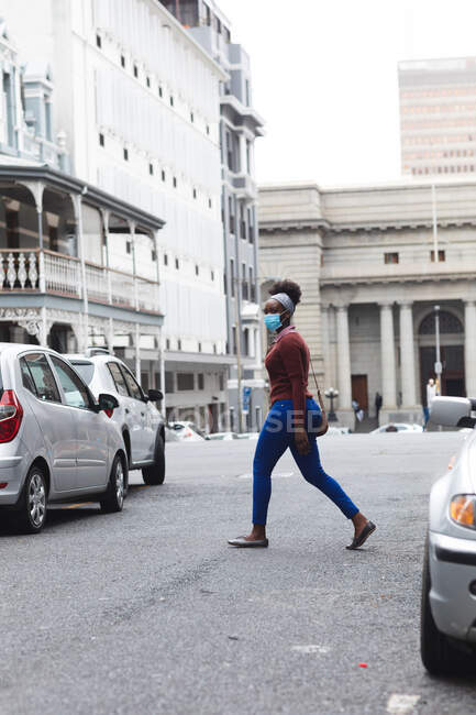 Femme afro-américaine portant un masque facial marchant dans la rue. dehors et autour dans la ville pendant la pandémie de coronavirus covid 19. — Photo de stock