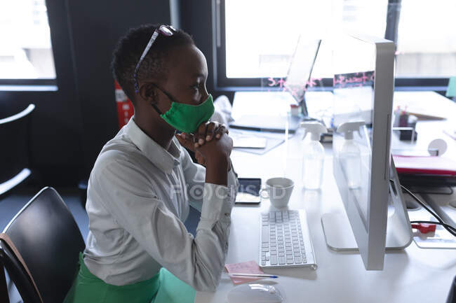 Африканська американка, одягнена в маску обличчя, дивиться на екран свого комп'ютера в сучасному офісі. Відстань до карантину під час пандемії коронавірусу — стокове фото