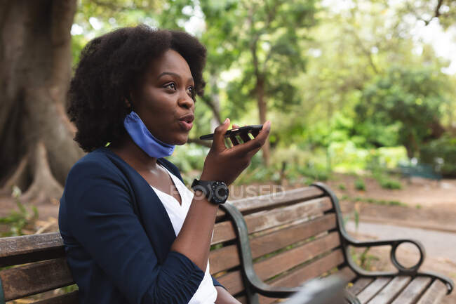 Mujer afroamericana con mascarilla en la calle sentada en un banco, hablando por teléfono. fuera de la ciudad durante la pandemia de coronavirus covid 19. - foto de stock