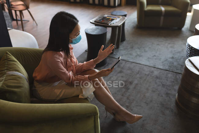 Mulher asiática usando máscara facial tendo um bate-papo por vídeo no smartphone no escritório moderno. bloqueio de quarentena por distanciamento social durante a pandemia do coronavírus — Fotografia de Stock