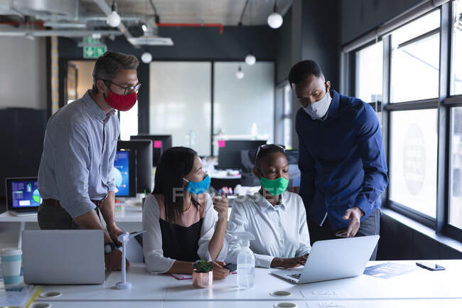 Разнообразные коллеги в масках для лица с помощью ноутбука во время совместной работы на современной офисной гигиене и социальной дистанцированности на рабочем месте во время пандемии коронавируса. — стоковое фото