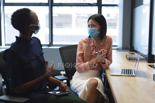 African american woman and asian woman wearing face masks talking to each other at modern office. social distancing quarantine lockdown during coronavirus pandemic — Stock Photo