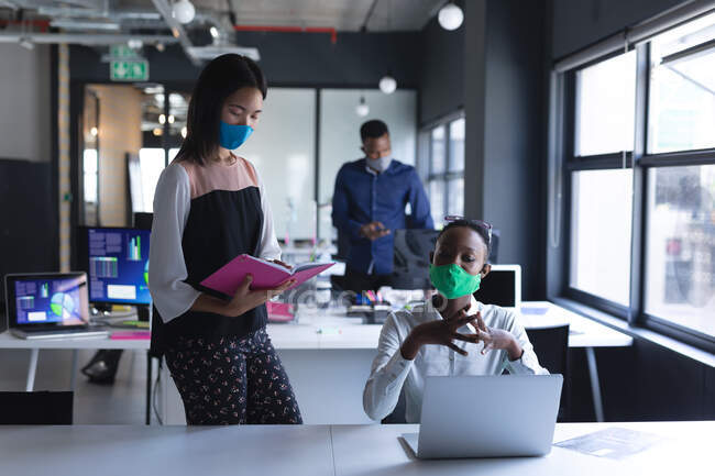 Asian woman reading book while african american woman using laptop at modern office. hygiene and social distancing in the workplace during coronavirus covid 19 pandemic. — Stock Photo
