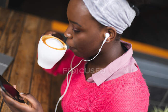 Donna afroamericana seduta in un caffè usando uno smartphone, bevendo una tazza di caffè e ascoltando musica. in giro per la città durante covid 19 coronavirus pandemia. — Foto stock