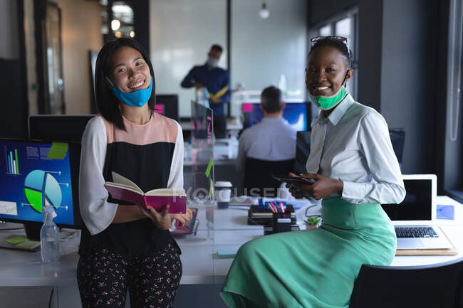 Portrait of asian woman with book and african american woman with digital tablet at modern office, social distancing quarantine lockdown during coronavirus pandemic — Stock Photo