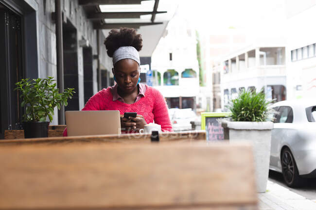 Африканская американка, сидящая в кафе на ноутбуке и смартфоне в городе во время пандемии коронавируса ковида 19. — стоковое фото