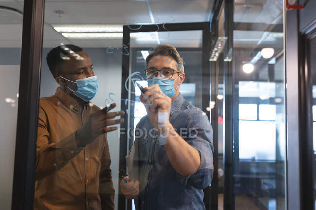 Homme caucasien portant un masque facial écrit avec un stylo marqueur sur un panneau de verre au bureau moderne. isolement social mise en quarantaine pendant une pandémie de coronavirus — Photo de stock