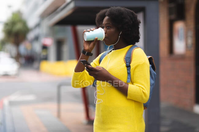 Mujer afroamericana usando smartphone en una calle bebiendo una taza de café y escuchando música con auriculares. fuera de la ciudad durante la pandemia de coronavirus covid 19. - foto de stock