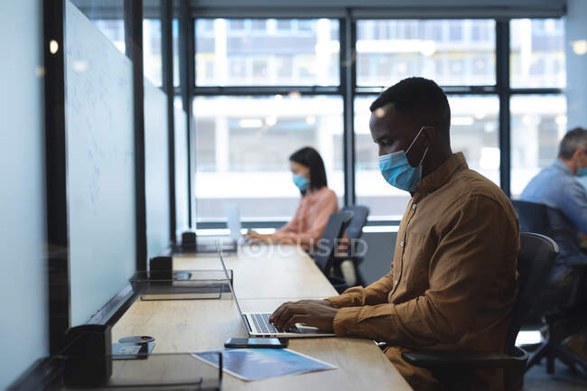 Afrikanischer Mann mit Mundschutz und Laptop, während er auf seinem Schreibtisch im modernen Büro sitzt. Soziale Distanzierung von Quarantäne während der Coronavirus-Pandemie — Stockfoto