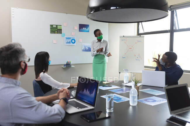 Африканская американка в маске для лица делает презентацию своим коллегам в современном офисе. гигиена и социальное дистанцирование на рабочем месте во время пандемии коронавируса 19 — стоковое фото