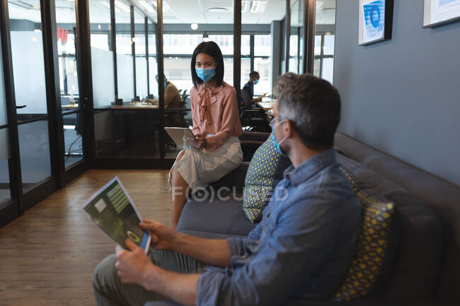 Asiatin trägt Gesichtsmaske mit digitalem Tablet, während kaukasischer Mann Dokumente im modernen Büro hält. Soziale Distanzierung von Quarantäne während der Coronavirus-Pandemie — Stockfoto