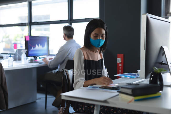 Азиатка в маске с графическим планшетом сидит на столе в современном офисе. гигиена и социальное дистанцирование на рабочем месте во время пандемии коронавируса 19. — стоковое фото